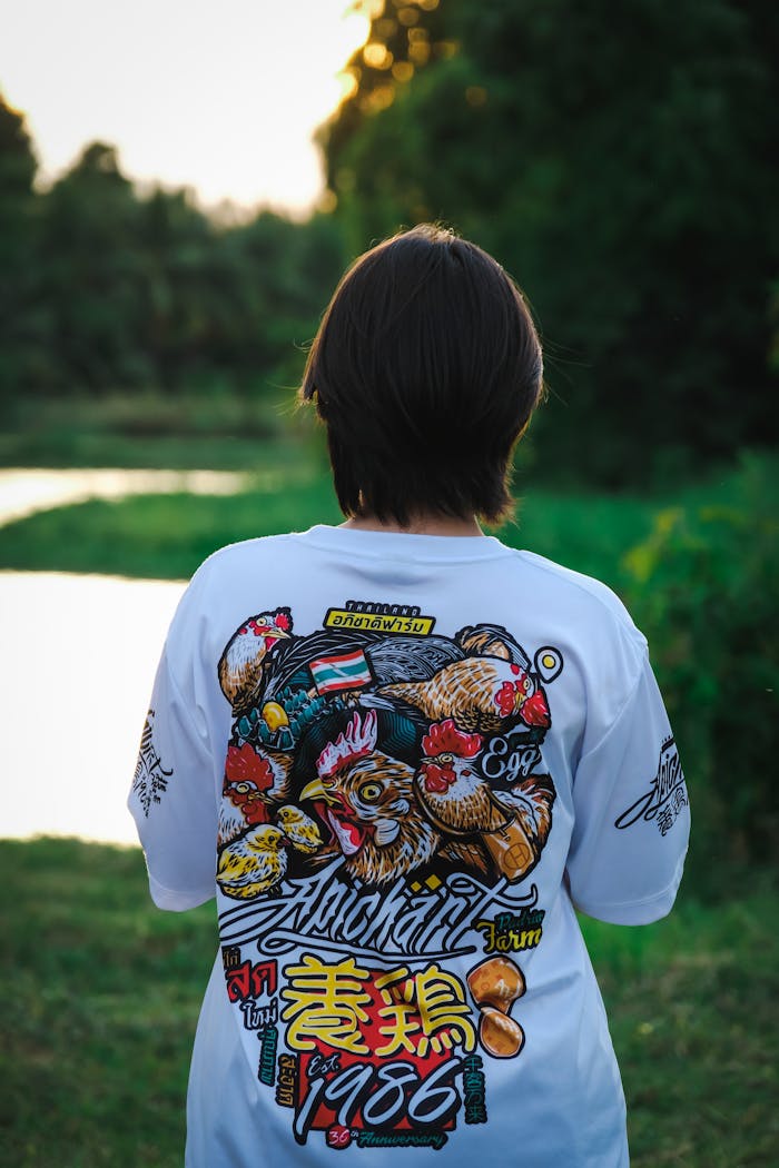 A young person with a graphic t-shirt stands near a tranquil lake in a forest setting.
