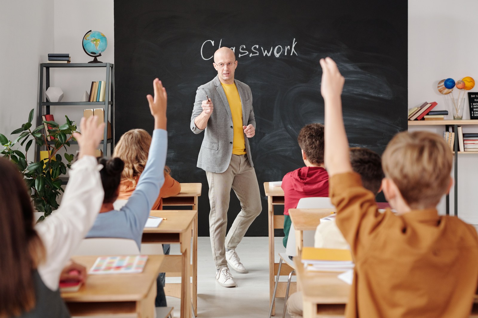 Dia do Professor: 4 educadores que mudaram o mundo Professora interagindo com alunos que levantam as mãos em sala de aula no Dia do Professor